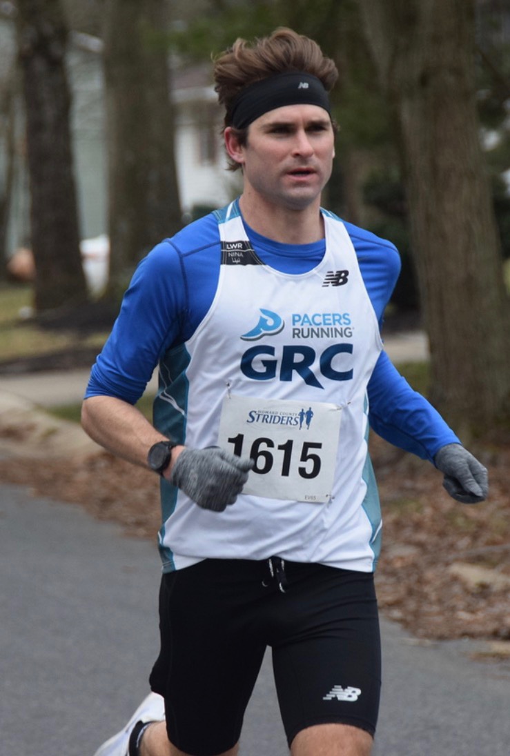 Dr. Christopher Bourke running a road race in a white and blue GRC Pacers Running singlet, race bib 1615
